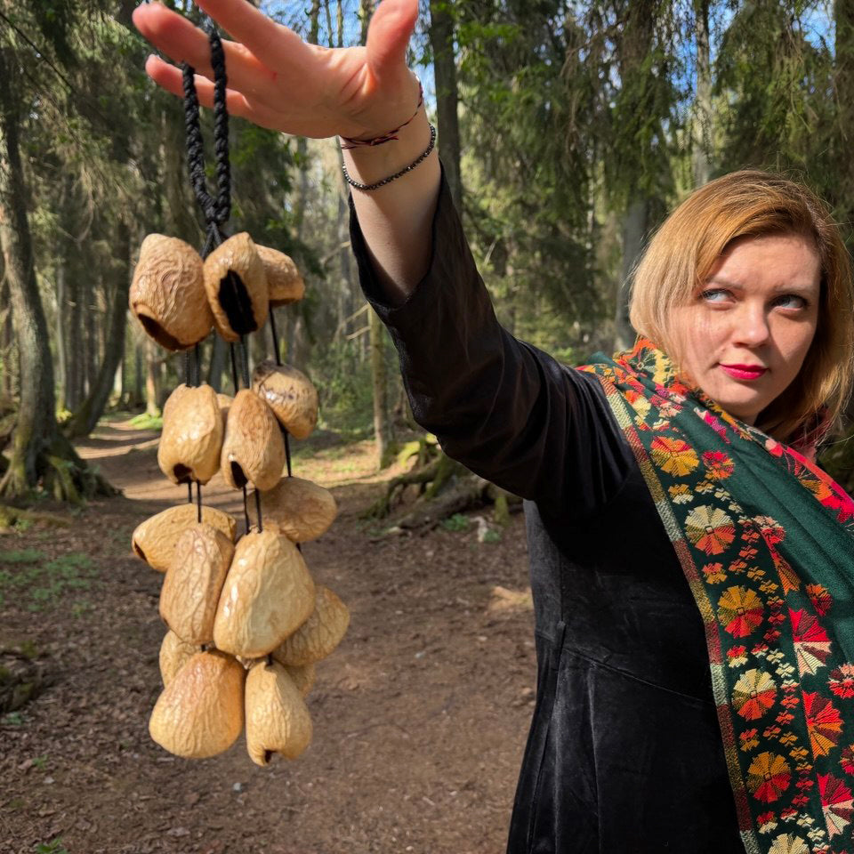 A woman in a forest setting holds up a cluster of natural wood cow bells on a black cord. The woman has short blonde hair and is wearing a black top and a colorful floral-patterned green scarf.