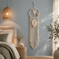 A cream macrame dreamcatcher hangs on a dusty blue wall in a styled bedroom setting, featuring a woven wicker pendant light, a wooden nightstand with a lit candle, and a bed with neutral-toned bedding.
