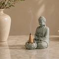 Ceramic Buddha statue with incense cone on a wooden surface, next to a vase.